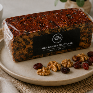 A packaged loaf of Rich Brandy Fruit Cake from The Original Cake Co, displayed on a beige ceramic plate with walnuts and dried cranberries. The cake is wrapped in clear plastic with a black label, set against a neutral, softly blurred background.