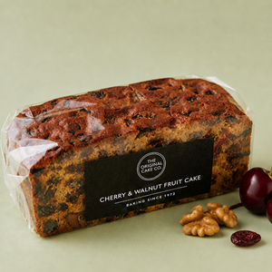 A packaged loaf of fruit cake on a light sage green background, with a few whole walnuts and dried cherries placed beside it. The cake is wrapped in clear plastic and has a black label reading 'The Original Cake Co.