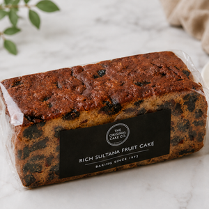 A loaf of Rich Sultana Fruit Cake from The Original Cake Co, wrapped in clear plastic with a black label, set on a light marble surface.