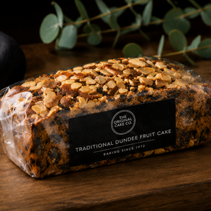 A packaged traditional Dundee fruit cake topped with sliced almonds and chopped nuts, wrapped in clear plastic with a black label reading 'The Original Cake Co.' and 'Traditional Dundee Fruit Cake,' set on a wooden surface with blurred greenery in the background.