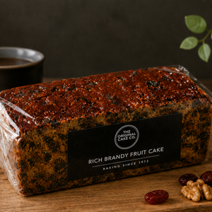 A loaf of Rich Brandy Fruit Cake by The Original Cake Co. wrapped in clear plastic with a black label, displayed on a wooden surface with dried fruits and nuts nearby.