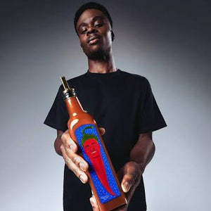 BUKAYO SAKA in a black t-shirt holds a brown glass bottle of chili oil with a colorful illustration of a red chili pepper on the label, set against a dark gray studio background.