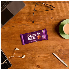 Cadbury Dairy Milk chocolate bar on a wooden desk with a laptop, glasses, earphones, and a cup.