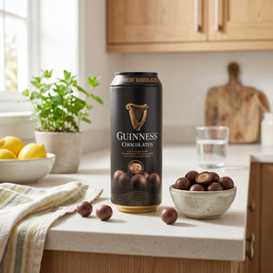 A can of Guinness Chocolates stands on a kitchen counter beside a bowl of chocolate-covered nuts, with lemons, a plant, and wooden cabinets in the background.