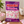 A bag of Haribo Chamallows Pink & White share size marshmallows stands on a wooden table, with pink and white marshmallows scattered around it. A bowl of marshmallows, a potted plant, and a cup of coffee are visible in the background.