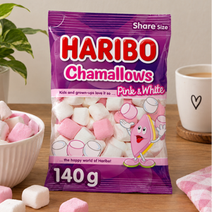 A bag of Haribo Chamallows Pink & White share size marshmallows stands on a wooden table, with pink and white marshmallows scattered around it. A bowl of marshmallows, a potted plant, and a cup of coffee are visible in the background.