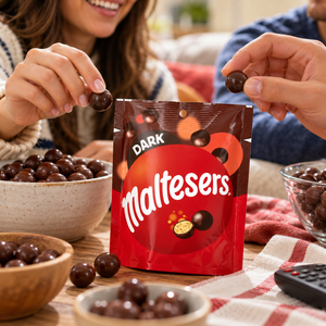A red bag of Maltesers Dark chocolate malt balls sits on a table, with bowls of the chocolate-covered treats and people reaching in from both sides.