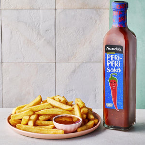 A bottle of Nando's Peri-Peri Sâka sauce stands next to a plate of french fries and a small dipping sauce dish on a light gray surface against a tiled wall.