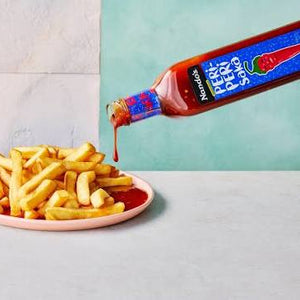 A bottle of Horseradish Pepper Sauce being poured onto a plate of french fries on a light gray surface, with a split background of white tiles and a mint green wall.