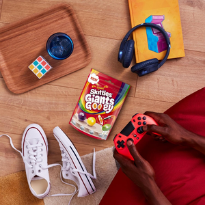 Person playing video games with a red controller, surrounded by snacks, shoes, and headphones on a wooden table.