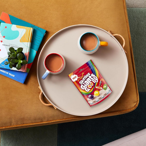 Round tray with two cups, a book, and a packet of sweets on a brown sofa.
