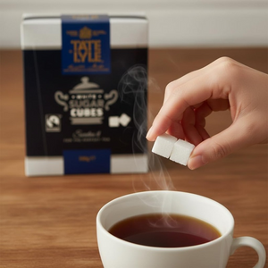 Person adding Tate & Lyle sugar cubes to a steaming cup of tea on a wooden surface.