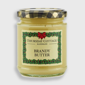 A glass jar of Thursday Cottage Handmade Brandy Butter with a black and gold checkered lid and a green holiday-themed label, set against a light gray background.