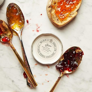 Two spoons with jam on a marble surface next to a jar lid and a piece of toast with jam.