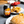 A can of Tango Orange Original soda placed on a gray tabletop next to plates of food, including a burger, fries, and fried chicken, with a red container in the background.