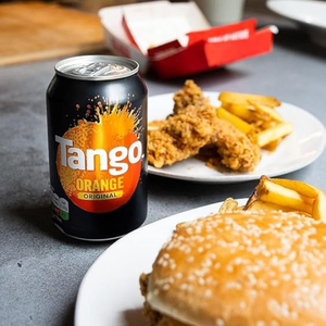 A can of Tango Orange Original soda placed on a gray tabletop next to plates of food, including a burger, fries, and fried chicken, with a red container in the background.