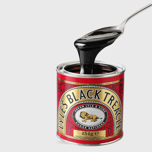 A tin of Lyles Black Treacle with a spoon lifting thick, glossy black syrup above the open container, on a light gray background.