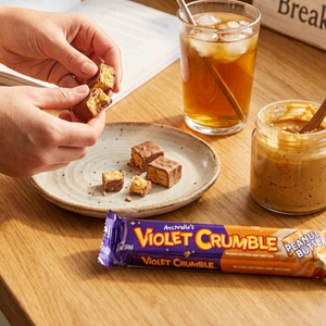 Person holding a Violet Crumble bar with a plate of brownies, iced tea, and peanut butter on a wooden table.