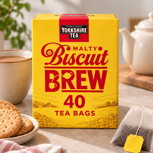 A yellow box of Yorkshire Tea Malty Biscuit Brew tea bags stands on a tabletop with a teapot, cookies, and a tea bag nearby, suggesting a cozy tea-time setting.