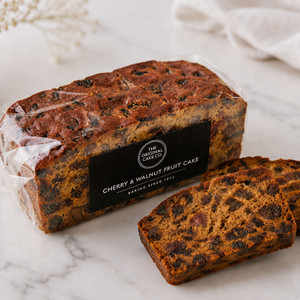 A loaf of cherry and walnut fruit cake in clear plastic packaging with a black label, alongside two sliced pieces showing the dense fruit pieces, set on a white marble surface.