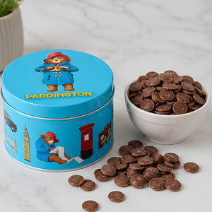 A blue Paddington bear metal tin filled with chocolate discs, shown beside a white bowl of more chocolates and scattered chocolate pieces on a light marble surface.