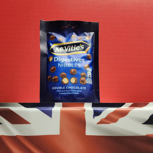 A package of McVitie's Digestives Nibbles Double Chocolate biscuit balls standing upright against a red background, with a Union Jack flag draped underneath the package.