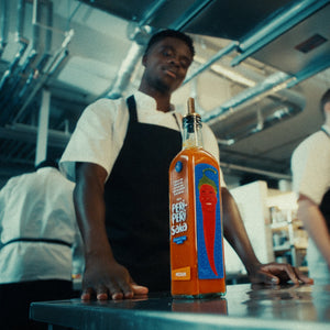 A bottle of Perch Fire Sauce stands on a stainless steel counter in a commercial kitchen, with two chefs in white shirts and black aprons blurred in the background.