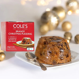 A box of Cole's Brandy Christmas Pudding stands next to a glazed Christmas pudding on a white plate with a spoon, set against a softly blurred festive background with gold ornaments and lights.